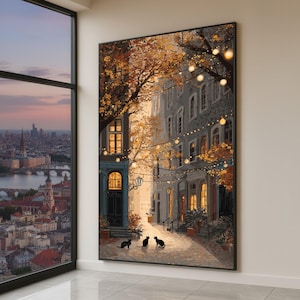 May include: A framed canvas print depicts a European street scene with buildings, trees, and string lights. Three black cats are in the foreground. The artwork features warm colors, creating a cozy, inviting atmosphere. The print is displayed on a white wall.