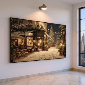 Può includere: Una stampa incorniciata raffigura una scena di strada innevata di notte. Un caffè con posti a sedere all'aperto è illuminato da luci calde e un albero di Natale si erge in lontananza. L'immagine è incorniciata in un bordo nero e appesa a una parete bianca.