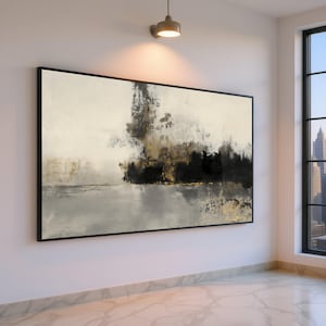 May include: Abstract art print in a black frame. The artwork features a blend of black, gold, and gray brushstrokes against a cream-colored background. The painting is displayed in a well-lit room with a city view.