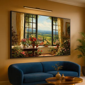 Op de afbeelding: Een groot ingelijst schilderij toont een schilderachtig uitzicht door een raam, met glooiende heuvels en een helderblauwe lucht. Het raam is versierd met bloemen en gordijnen, wat een warme sfeer creëert. Een blauwe bank en salontafel staan op de voorgrond, wat een gezellige woonruimte creëert.