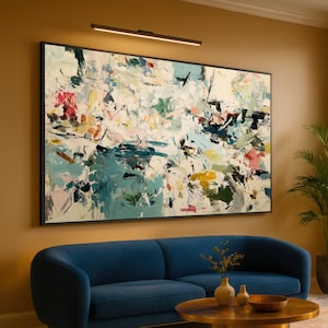 Könnte beinhalten: Ein abstraktes Gemälde in einem schwarzen Rahmen hängt über einem blauen Sofa. Das Kunstwerk zeigt eine Mischung aus Farben, darunter Türkis, Weiß, Rosa und Gelb. Über dem Gemälde ist eine moderne schwarze Leuchte angebracht. Ein goldener Couchtisch und dekorative Vasen befinden sich vor dem Sofa.