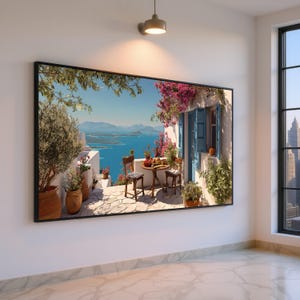 Puede incluir: Impresión enmarcada de una escena de balcón mediterráneo. La obra de arte presenta una mesa para dos, rodeada de flores y plantas, con contraventanas azules y una vista al mar y las montañas.