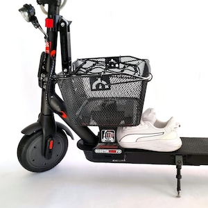 Op de afbeelding: Zwarte elektrische scooter met een zwarte draadmand en een witte sneaker. De scooter heeft een koplamp, een rode accentkleur en een zwarte band. De mand is op het stuur gemonteerd.