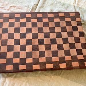 May include: A rectangular wooden chopping board featuring an end-grain construction with a repeating checkerboard pattern. The surface is composed of alternating light tan and dark brown wood squares, creating a high-contrast grid design.