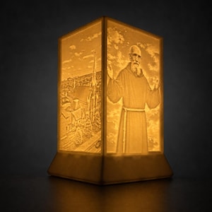Peut inclure: Une lampe lithophane rectangulaire diffusant une lueur ambrée chaude. Une face présente le relief détaillé d'un personnage barbu en robe à capuche, les bras levés. La face adjacente montre le relief d'une église avec un clocher et des arbres.