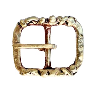 Puede incluir: Una hebilla de cinturón de metal dorado con un diseño trenzado. La hebilla es rectangular con un borde redondeado y una sola abertura rectangular en el centro.