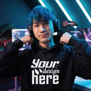 Könnte beinhalten: Ein schwarzer Kapuzenpullover mit dem weißen Text "Your design here". Die Person auf dem Bild trägt Kopfhörer und hat blaue Haare. Der Hintergrund ist ein Gaming-Setup mit Monitoren und Neonlichtern.