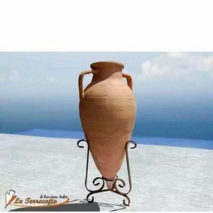 May include: A terracotta amphora, a large, ancient Greek jar, stands on a black metal stand. The amphora is a traditional pottery vessel with a rounded body and a narrow neck. It is displayed against a backdrop of a blue sky and ocean.