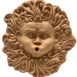 Peut inclure: Une sculpture murale en terre cuite représentant un visage avec un vent stylisé qui souffle autour de lui. Le visage a une large bouche avec un trou au centre, et le vent est composé de lignes tourbillonnantes.