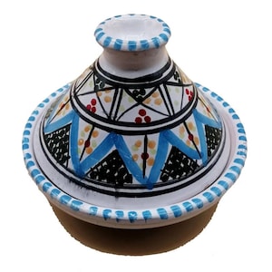 Puede incluir: Una olla de tagine de cerámica blanca con tapa cónica. La olla presenta un patrón geométrico detallado en negro, azul y rojo. El borde y la tapa están adornados con detalles azules, creando un artículo de cocina vibrante y decorativo.
