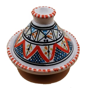Puede incluir: Una olla de tagine de cerámica con base y tapa blancas, adornada con un colorido patrón geométrico en rojo, azul, naranja y negro. El borde de la tapa y la base presenta un diseño punteado rojo. La olla está diseñada para cocinar y servir.