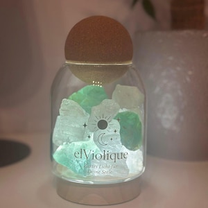 Könnte beinhalten: Ein Glasbehälter mit Korkdeckel, gefüllt mit grünen und weißen Kristallen. Auf dem Glas steht "elViolique" und "Zartes Licht für Deine Seele". Der Behälter steht auf einem Holzsockel.