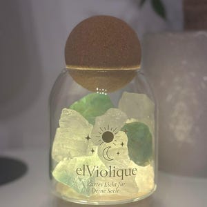 Könnte beinhalten: Ein Glasbehälter mit Korkdeckel, gefüllt mit leuchtenden grünen und weißen Kristallen. Der Behälter hat einen hellen Sockel und trägt den Text "elViolique" mit einem Sonnen- und Monddesign. Der Text "Zartes Licht für Deine Seele" ist ebenfalls sichtbar.