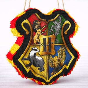 Könnte beinhalten: Eine schildförmige Piñata mit dem Wappen von Hogwarts. Das Wappen ist in vier Abschnitte unterteilt, jeder mit einem anderen Tier und einer anderen Farbe. Die Piñata hat einen schwarzen Rand mit roten und gelben Fransen und ein Seil zum Aufhängen.