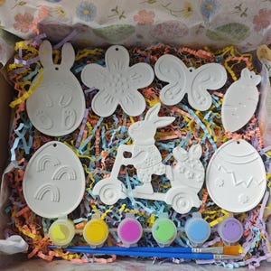 Può includere: Un kit artigianale a tema pasquale con varie forme in gesso bianco, tra cui un coniglietto, un fiore, una farfalla, una carota e uova di Pasqua. Il kit include vasetti di vernice multicolore e pennelli, il tutto in una scatola decorativa.
