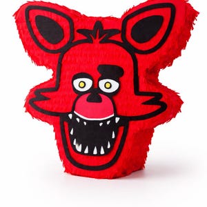 May include: A red, plush piñata in the shape of a cartoon fox head. The fox has black outlines, ears, and a wide, toothy grin. The eyes are white with yellow pupils. The piñata is designed for a party or celebration.