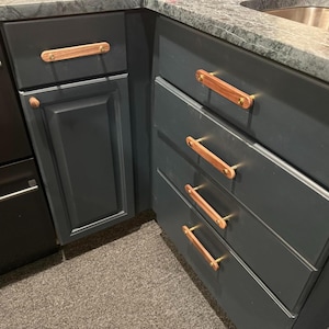 May include: Dark grey kitchen cabinets with wooden and brass handles. The cabinets have a flat panel design and are arranged in a corner configuration. The wooden handles are rectangular and attached with brass screws.