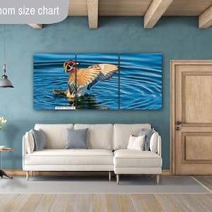 May include: A three-panel wall art print featuring a colorful duck with outstretched wings in a blue water setting. The artwork is displayed above a white sectional sofa with gray and white pillows. A wooden door and a small side table with flowers are also visible.