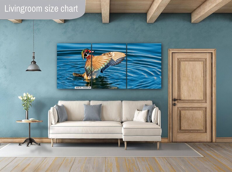 May include: A three-panel wall art print featuring a colorful duck with outstretched wings in a blue water setting. The artwork is displayed above a white sectional sofa with gray and white pillows. A wooden door and a small side table with flowers are also visible.
