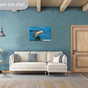 May include: A framed print of a bird in water, with wings spread, is displayed above a white sofa. The room has a teal wall, a wooden door, and a wooden ceiling. A side table with a lamp and a vase of flowers is in the foreground.