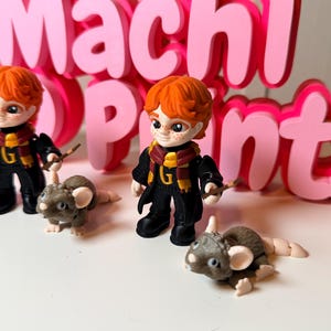 May include: Three figurines of a character with red hair, wearing a black robe, scarf, and holding a wand, accompanied by small gray mouse figurines. The background features a pink sign that reads "Machi Prints".