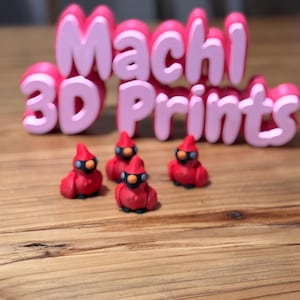 May include: Four red cardinal bird figurines with black eyes and beaks. The figurines are sitting on a wooden surface in front of a pink sign that says "Machi 3D Prints".