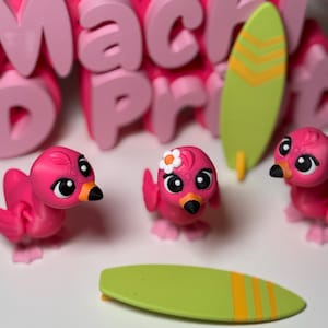May include: Three pink flamingo figurines with black eyes and orange beaks. One flamingo has a white flower on its head. The figurines are on a white surface with a green surfboard with yellow stripes.