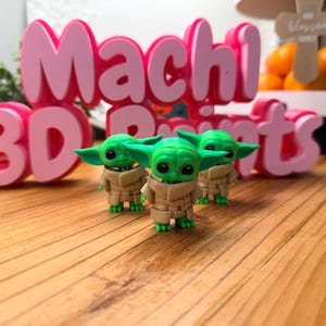 May include: Three green figurines of a character with large ears and a tan coat stand in front of pink 3D printed letters that spell "Machi 3D Prints". The figurines are on a wooden surface.