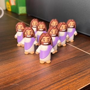 May include: A group of ten small, brown figurines of Jesus with purple robes and white tunics. The figurines are arranged in a semi-circle on a wooden surface.