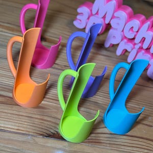 May include: A collection of colorful 3D-printed spoon rests in shades of pink, orange, green, purple, and blue. Each rest features a curved handle and a deep, rounded bowl. The text "3D Mach Print" is visible in the background.