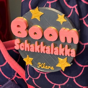 Könnte beinhalten: Ein rosa-goldenes 3D-gedrucktes Namensschild mit dem Text "Boom Schakkalakka Klara" auf grauem Hintergrund.