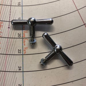 Pode incluir: Dois botões cromados em forma de T, cada um com uma esfera central e três braços, fixados a parafusos com arruelas. Os botões estão sobre uma superfície com um padrão circular e o texto "lace Hoop Seam Here".