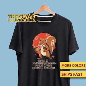 May include: Black t-shirt featuring a cartoon squirrel holding acorns, set against a red circle with falling leaves. The text "EVEN A BLIND SQUIRREL FINDS A NUT ONCE IN A WHILE" is below the graphic. The shirt is hanging on a wooden hanger.