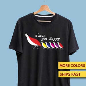 Peut inclure: T-shirt noir avec un motif d'oiseaux stylisés rouges, jaunes, bleus et violets, et le texte "c'mon get happy". Le t-shirt est suspendu à un cintre en bois sur fond bleu clair.
