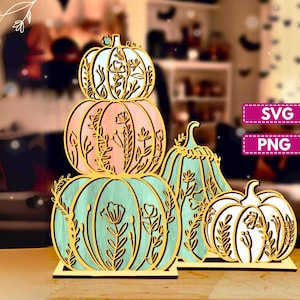 May include: Wooden pumpkin decorations in various sizes and colors. The pumpkins are stacked and arranged, featuring intricate floral cutouts. The color palette includes shades of pink, teal, and white, with gold accents. The image also includes the text "SVG" and "PNG".