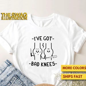 Könnte beinhalten: Weißes T-Shirt mit einer schwarzen Grafik von zwei Kniegelenken, eines mit einem Messer und das andere mit einem Baseballschläger, mit dem Text "-I'VE GOT- BAD KNEES-". Das Logo The Swag Illustration befindet sich oben links.