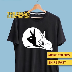 May include: Black t-shirt featuring a white rabbit and a hand shadow forming a rabbit shape. The text "THE SWAG ILLUSTRATION" is above the graphic. The shirt is displayed on a wooden hanger.