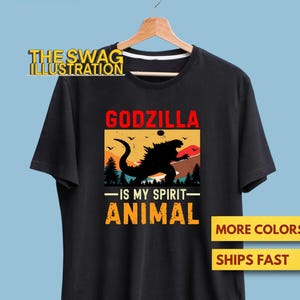 May include: Black t-shirt featuring a Godzilla graphic. The design includes the word "GODZILLA" in red, a black silhouette of Godzilla, and the text "-IS MY SPIRIT- ANIMAL" in orange. The shirt is hanging on a wooden hanger.
