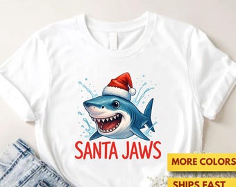 May include: White t-shirt featuring a cartoon shark wearing a Santa hat, with the text "SANTA JAWS" in red. The shark is blue with white teeth and water splashes around it. The shirt is displayed with denim shorts.