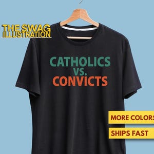 Puede incluir: Camiseta negra con el texto "CATHOLICS VS. CONVICTS" en verde y naranja. La camiseta está colgada de una percha de madera sobre un fondo azul claro. El logotipo de The Swag Illustration es visible.