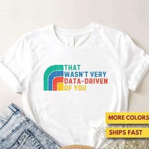 May include: White t-shirt featuring the phrase "THAT WASN'T VERY DATA-DRIVEN OF YOU" in a retro font with a rainbow design. The shirt is paired with denim jeans. Additional text on the image says "MORE COLORS" and "SHIPS FAST".