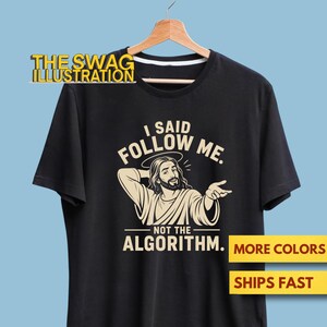 Może przedstawiać: Czarny t-shirt z krótkim rękawem z kremowym nadrukiem graficznym. Wzór przedstawia postać z aureolą oraz napis "I SAID FOLLOW ME. NOT THE ALGORITHM." Koszulka jest zawieszona na drewnianym wieszaku na jasnoniebieskim tle.