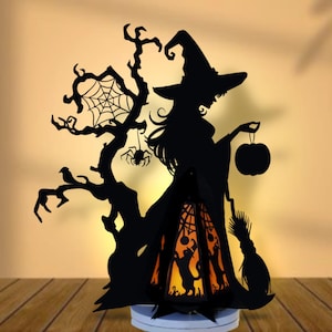 May include: Black silhouette Halloween decoration featuring a witch holding a pumpkin, a tree with a spider web, and a broom. The witch stands next to a lantern with a cat silhouette and an orange glow. The decoration is on a white base.