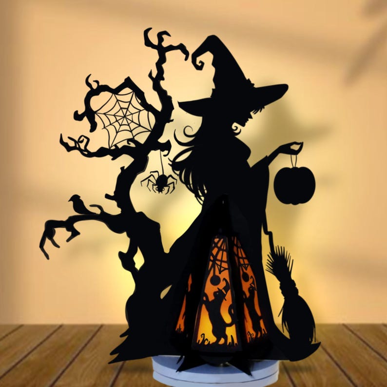 May include: Black silhouette Halloween decoration featuring a witch holding a pumpkin, a tree with a spider web, and a broom. The witch stands next to a lantern with a cat silhouette and an orange glow. The decoration is on a white base.