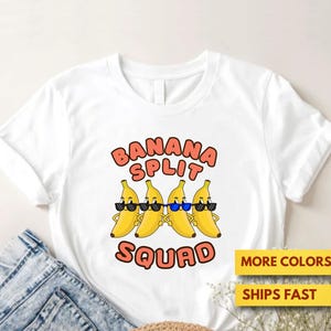 Puede incluir: Camiseta blanca con un gráfico de cuatro plátanos amarillos con gafas de sol y el texto "BANANA SPLIT SQUAD" en rojo y naranja. La camiseta se muestra con vaqueros.