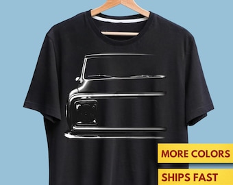 Puede incluir: Camiseta negra con una ilustración en escala de grises de la parte delantera de un camión clásico. El diseño incluye el capó, los faros y el parachoques. La camiseta cuelga de una percha de madera sobre un fondo azul claro. El texto en la camiseta dice "MORE COLORS" y "SHIPS FAST".