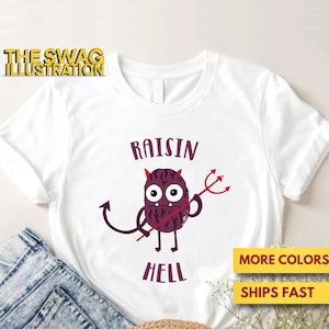 Puede incluir: Camiseta blanca con un diablo de pasa de dibujos animados sosteniendo un tridente y el texto "RAISIN HELL". El texto "THE SWAG ILLUSTRATION" está en la parte superior izquierda. La camiseta se muestra con vaqueros azules.