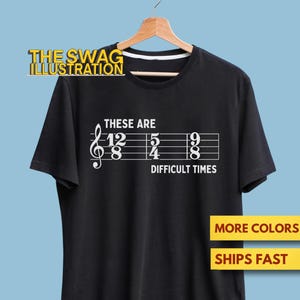 Può includere: T-shirt nero con un design a pentagramma bianco. Il design include una chiave di violino, note musicali e il testo "THESE ARE DIFFICULT TIMES". La t-shirt è esposta su una gruccia di legno su sfondo azzurro.
