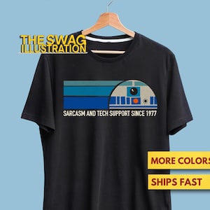 May include: Black t-shirt featuring a retro-style graphic of a droid with blue and teal stripes above it. The text "Sarcasm and Tech Support Since 1977" is printed below the graphic. The shirt is hanging on a wooden hanger.