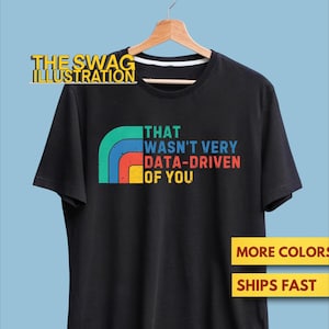May include: Black t-shirt with the text "THAT WASN'T VERY DATA-DRIVEN OF YOU" in a retro font. A rainbow design is above the text. The Swag Illustration logo is at the top left. The shirt is hanging on a wooden hanger.